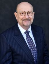 Portrait of a smiling man in a navy pinstripe suit, white shirt, patterned tie, and round glasses on a dark background