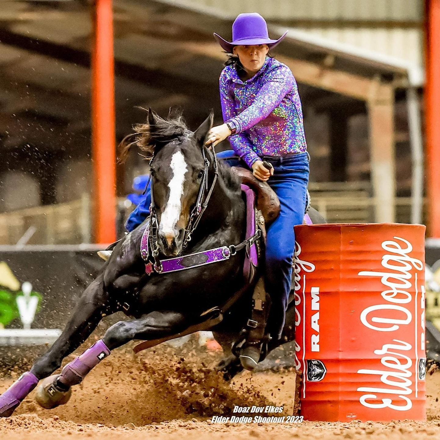 Cowgirl on a black horse racing around a red barrel, kicking up dirt in an arena.