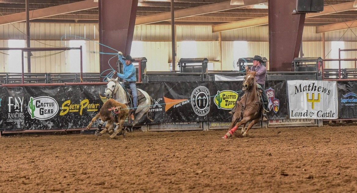 Two riders on horses race in a dirt arena during a rodeo, with sponsor banners in the background.