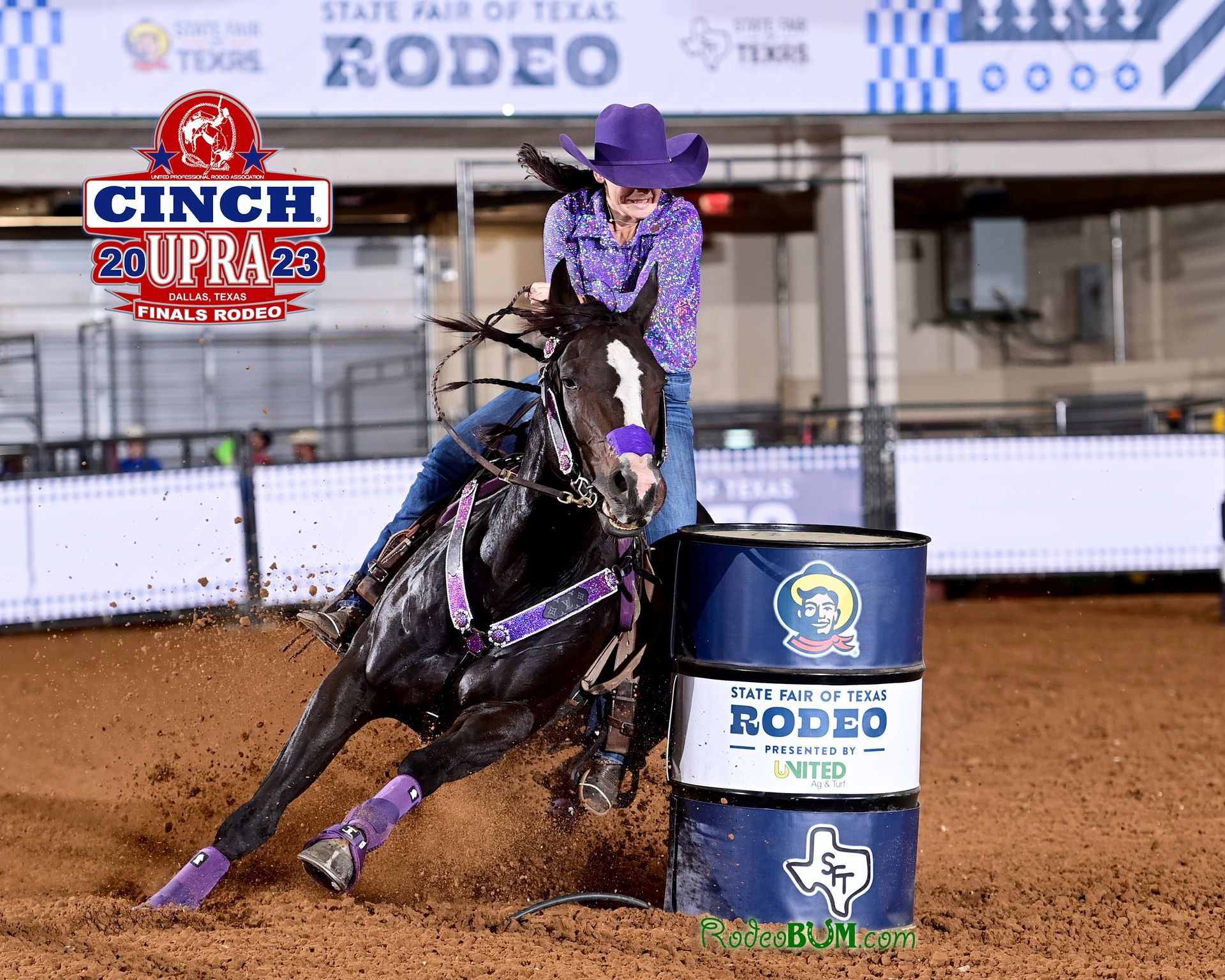 Barrel racer on a black horse leans hard around a turn in a rodeo arena, kicking up dirt.