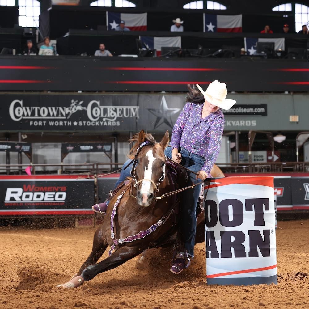 Rodeo rider in purple shirt leans hard around a barrel on a brown horse in an indoor arena.