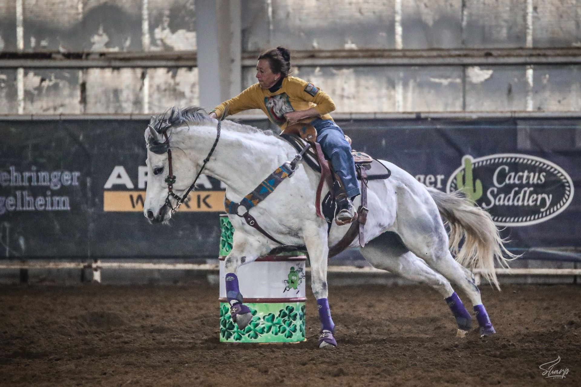 Cowboy on a gray horse racing around a barrel in an indoor arena, kicking up dust.