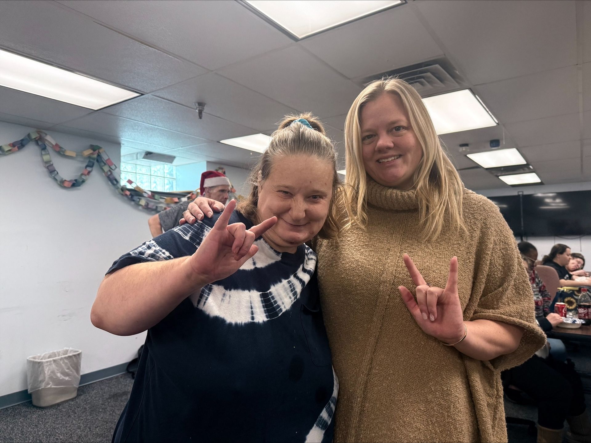 Two people smiling and posing indoors, one in a navy shirt and one in a beige sweater, flashing peace signs.