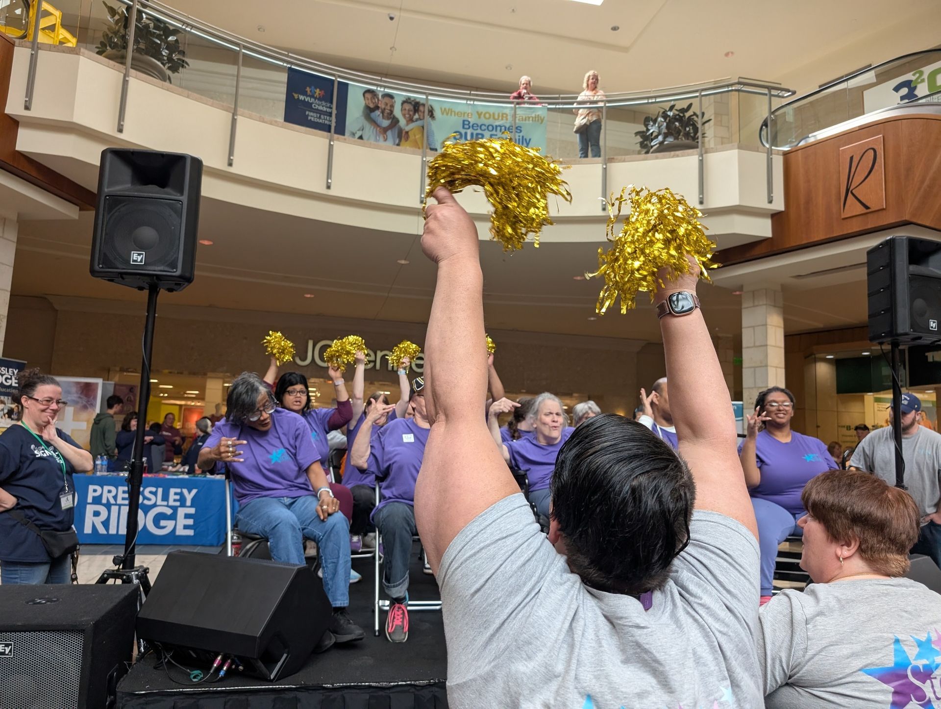 Group of people in purple shirts cheer with gold pom-poms at an indoor event.