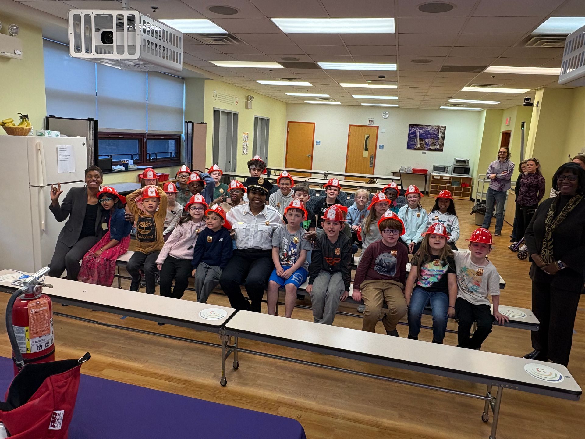 Group of children wearing red hats seated in a classroom or cafeteria, facing the camera and smiling.