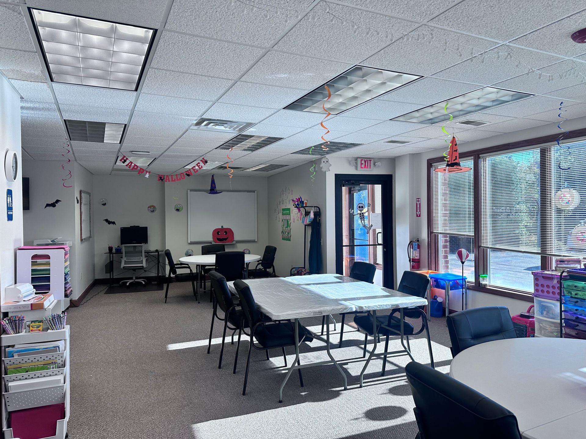 Brightly lit room with tables, chairs, whiteboard, and decorations, likely for a gathering or class.