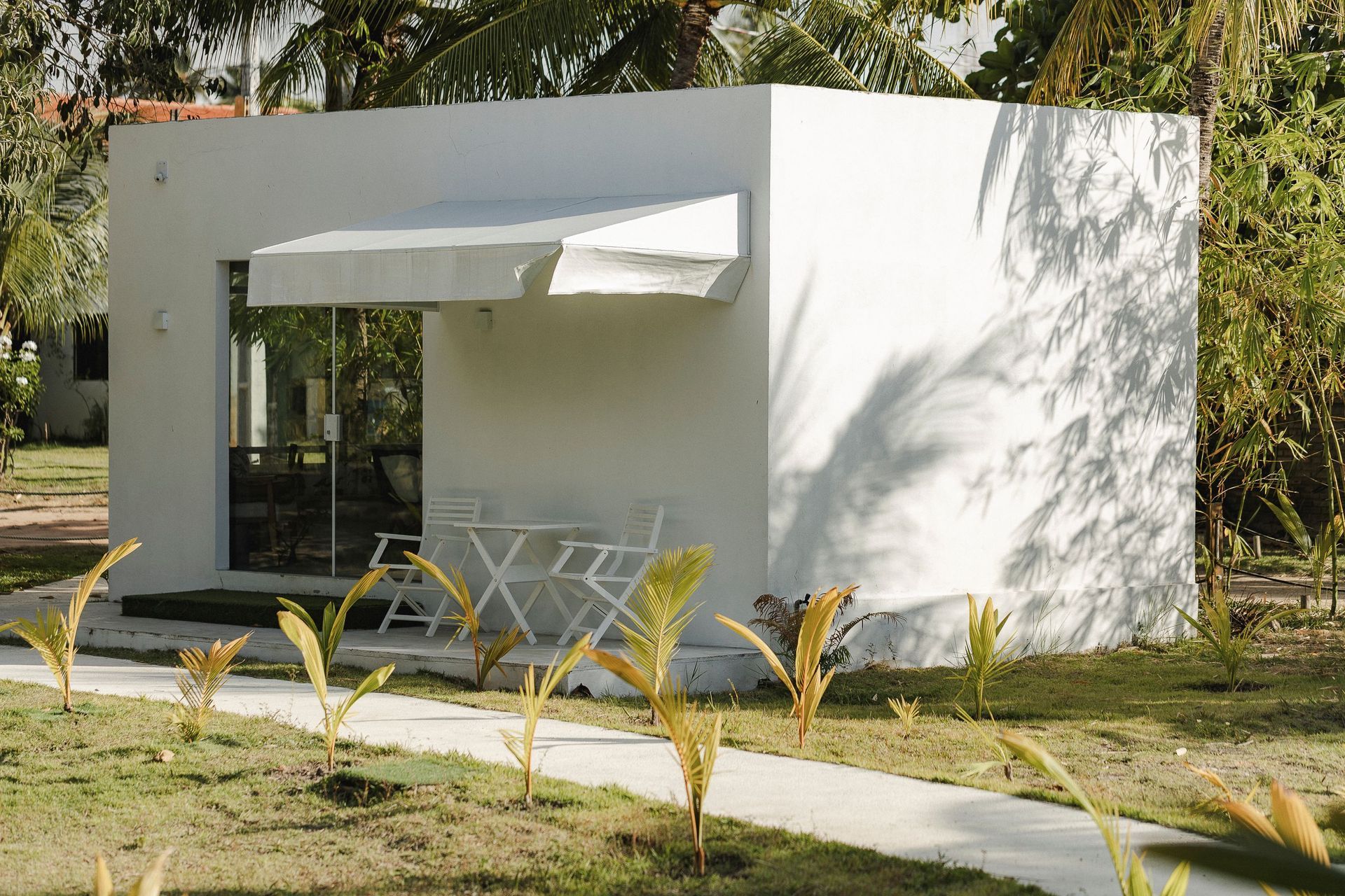 Pavilhão moderno branco para pátio com toldo, porta de vidro e mesa e cadeiras externas em um jardim.