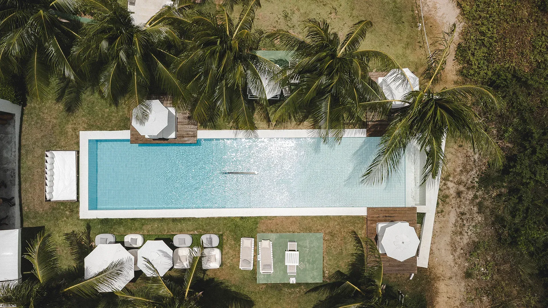 Vista aérea de uma piscina no quintal com espreguiçadeiras, palmeiras e uma área de estar sombreada.