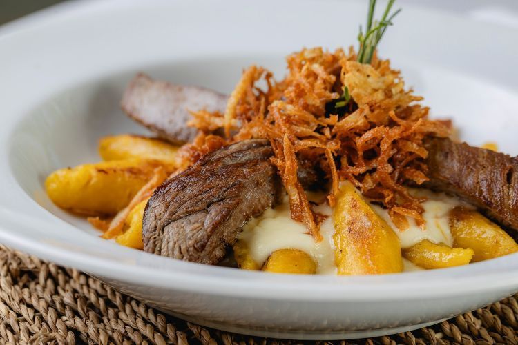 Bife servido com batatas fritas e cebolas crocantes em uma tigela branca, decorado com alecrim.