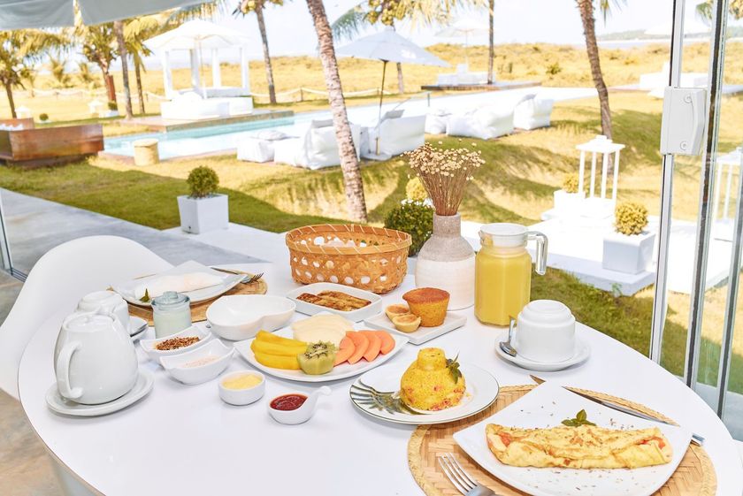 Café da manhã servido em uma mesa branca no pátio, com vista para uma piscina ensolarada e um resort repleto de palmeiras.