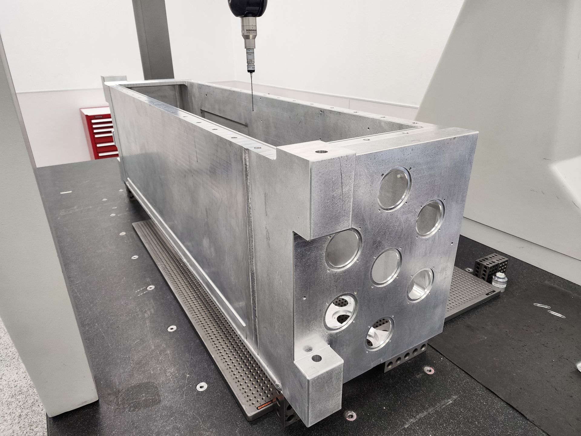 Metal rectangular part being measured by a CMM (Coordinate Measuring Machine) in a manufacturing setting.
