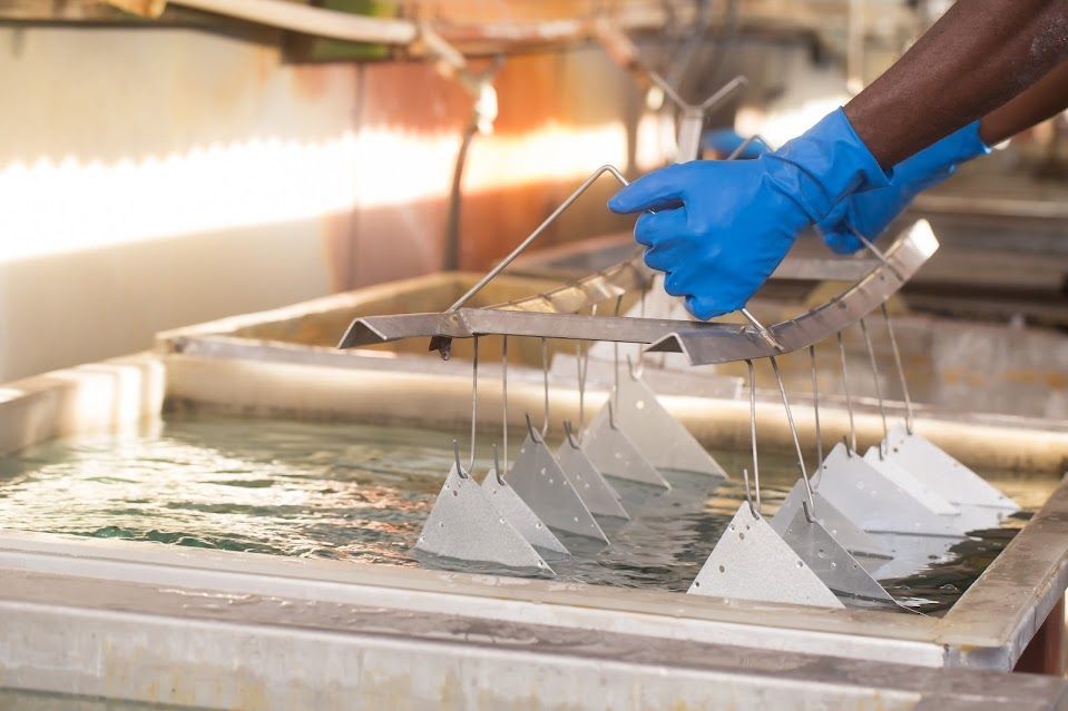 Person in blue gloves dips metal parts into a tank of liquid.