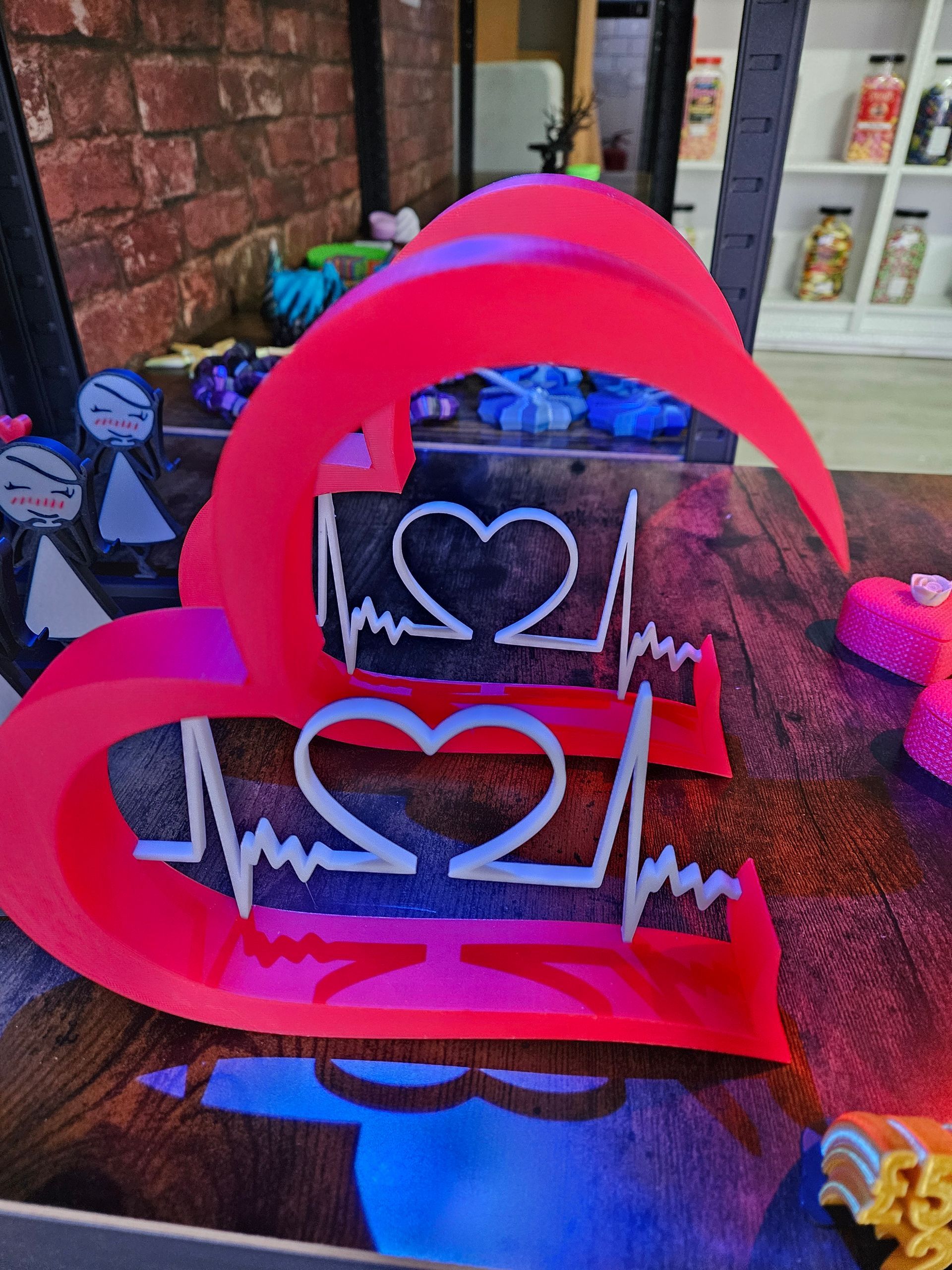 Pink heart-shaped sign with white heart and heartbeat design on a table. Brick wall background.