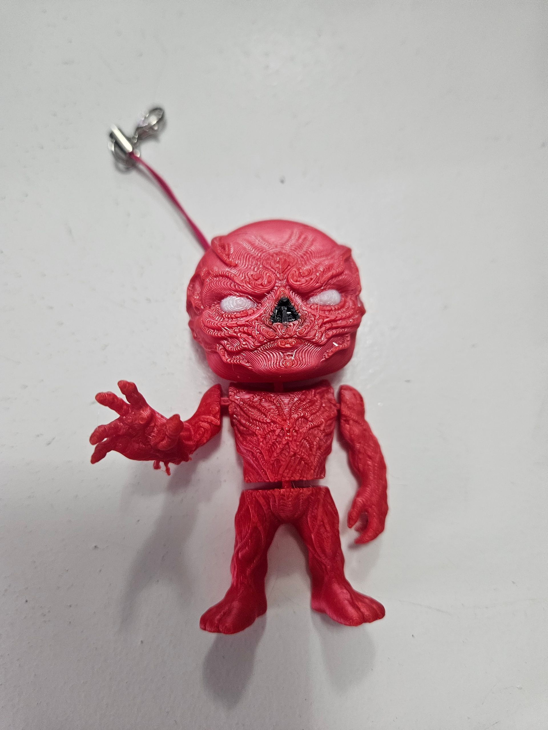 Red monster figurine with a string. It has a rough texture, white eyes, and outstretched arm.