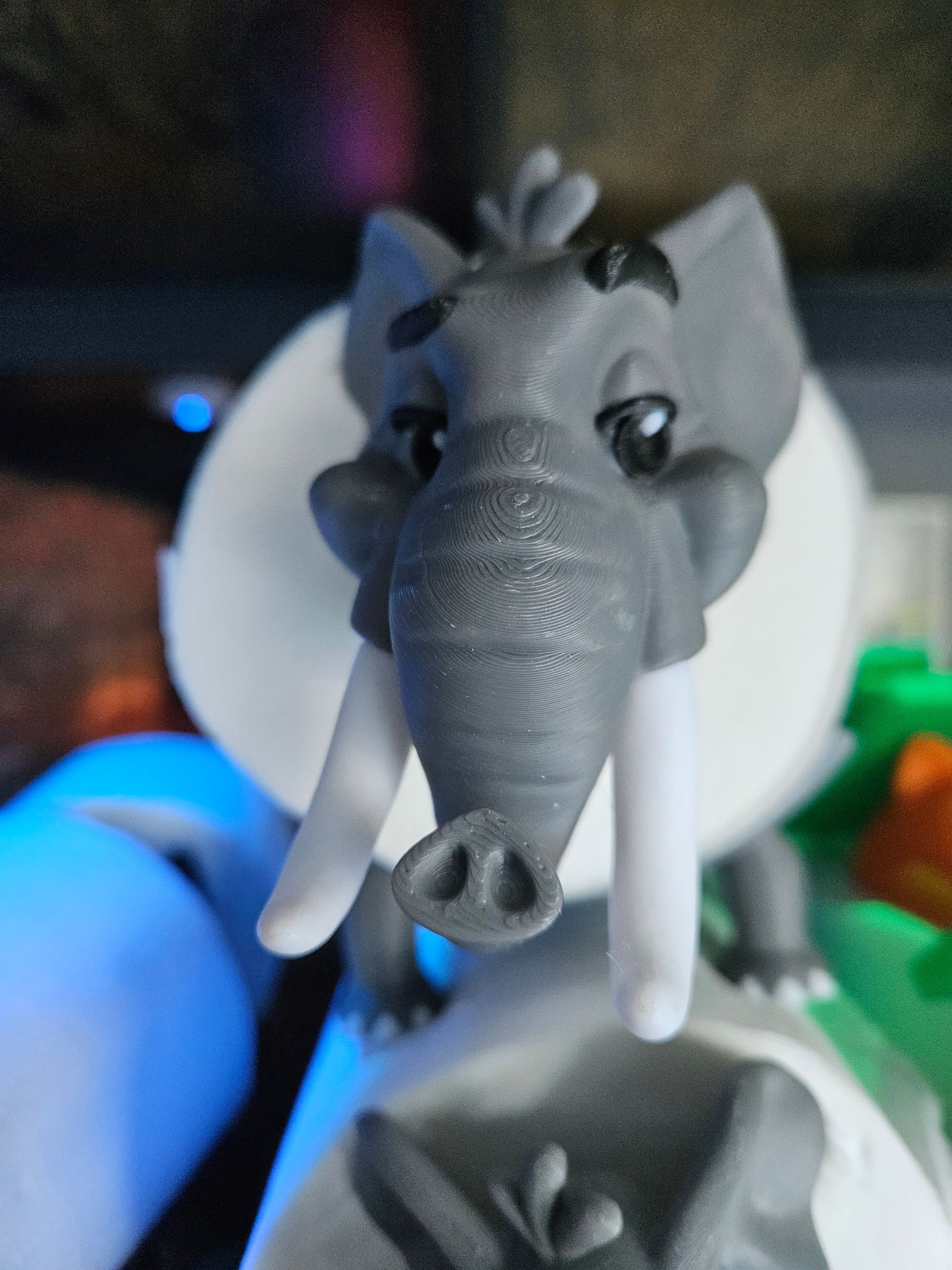 Gray toy elephant with tusks, front-facing close-up, against a blurred colorful background