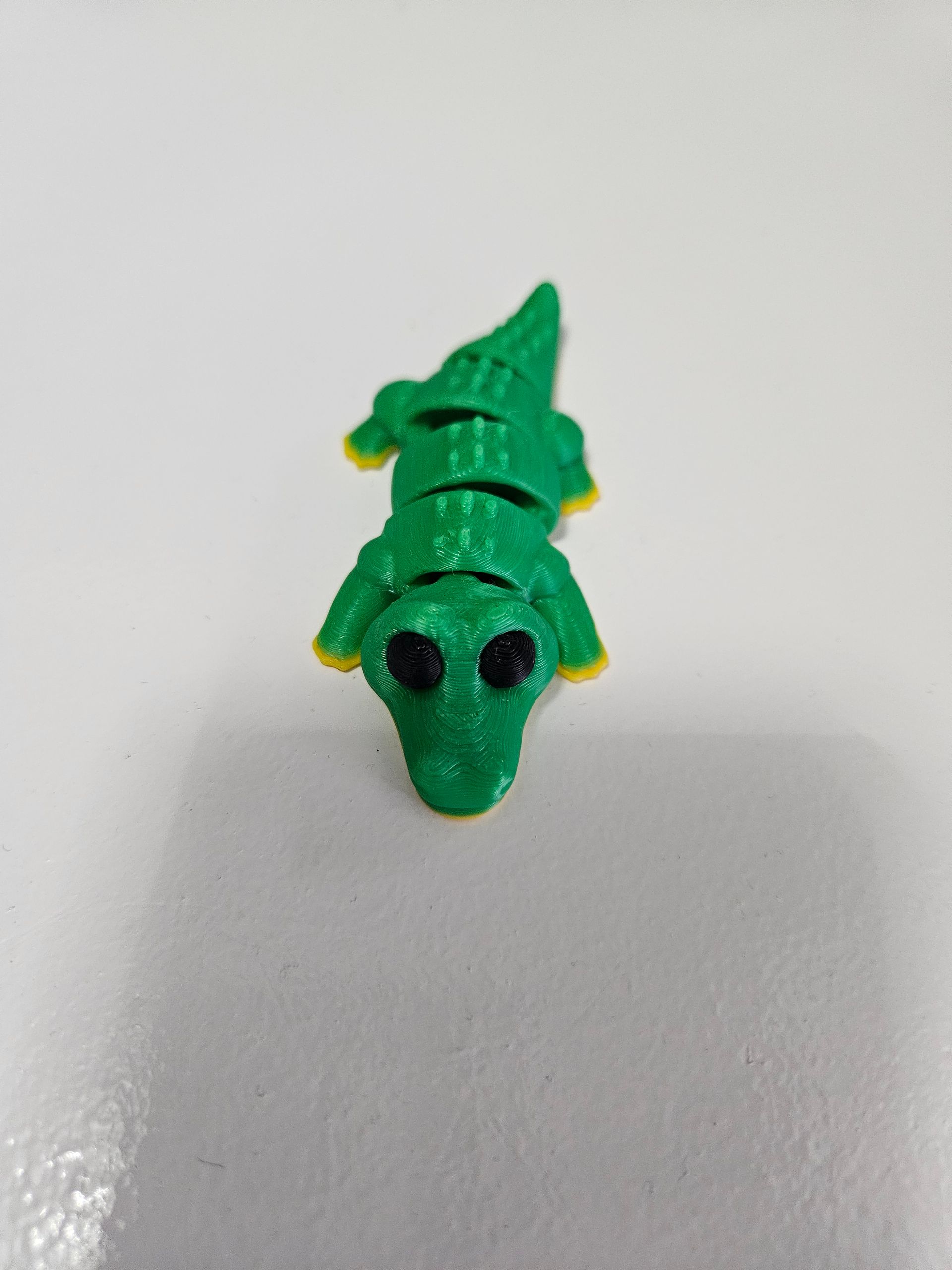 Green and yellow articulated toy alligator on a white surface, with black eyes.