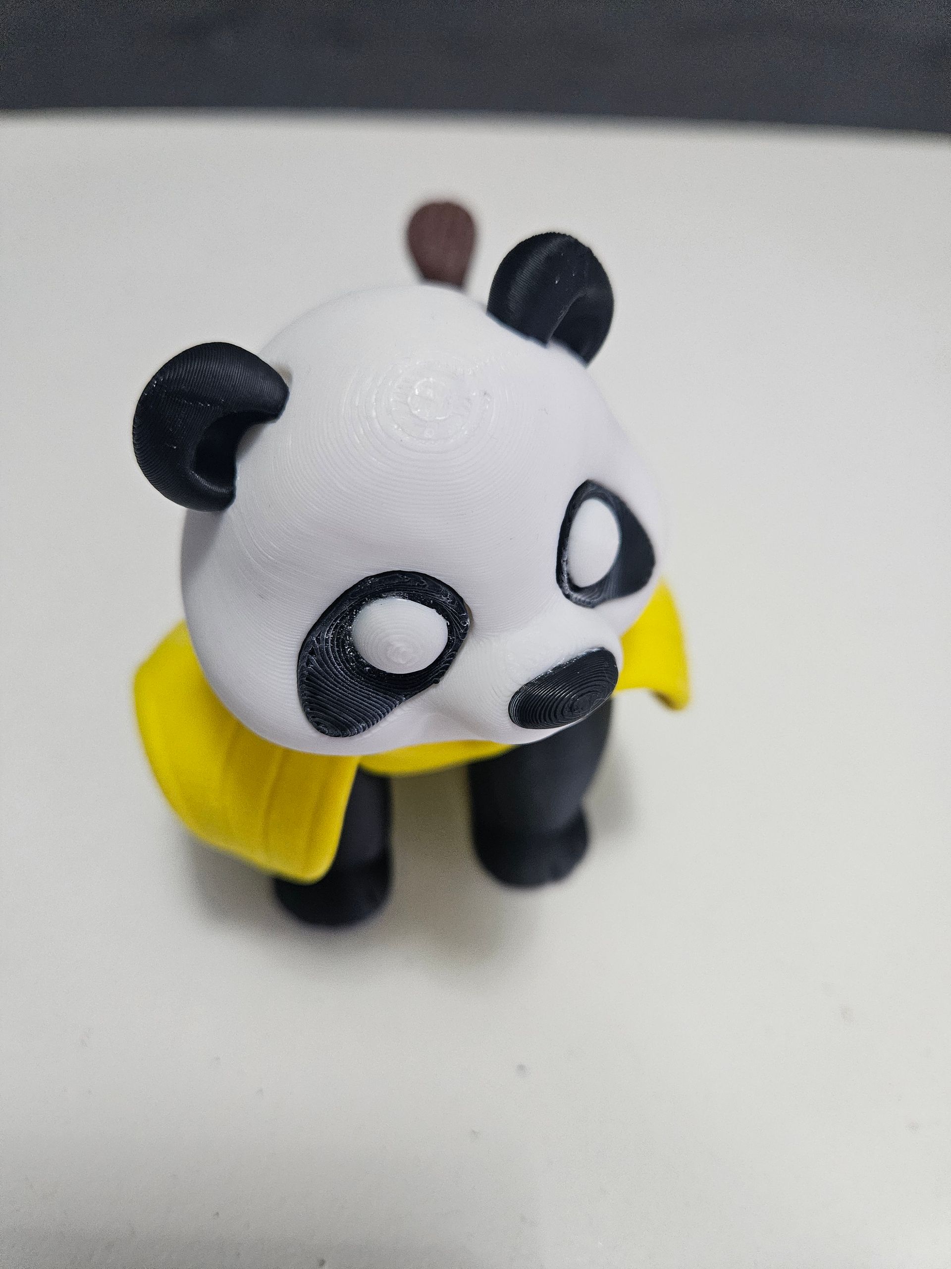 A small, 3D-printed panda with black and white fur, wearing a yellow cape, and brown stick on its back.