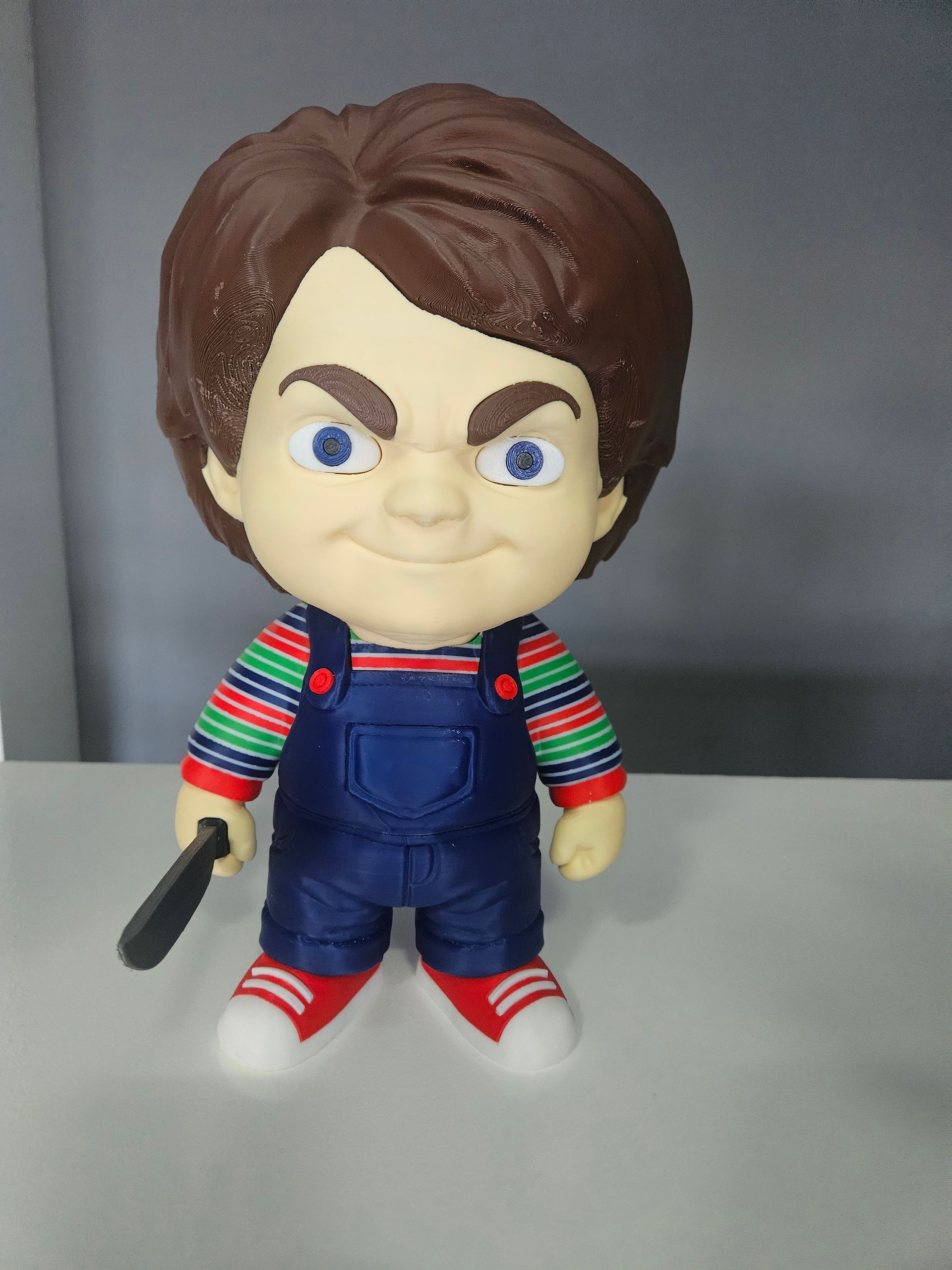 Chucky doll figurine with striped shirt, denim overalls, and a knife, standing against a gray background