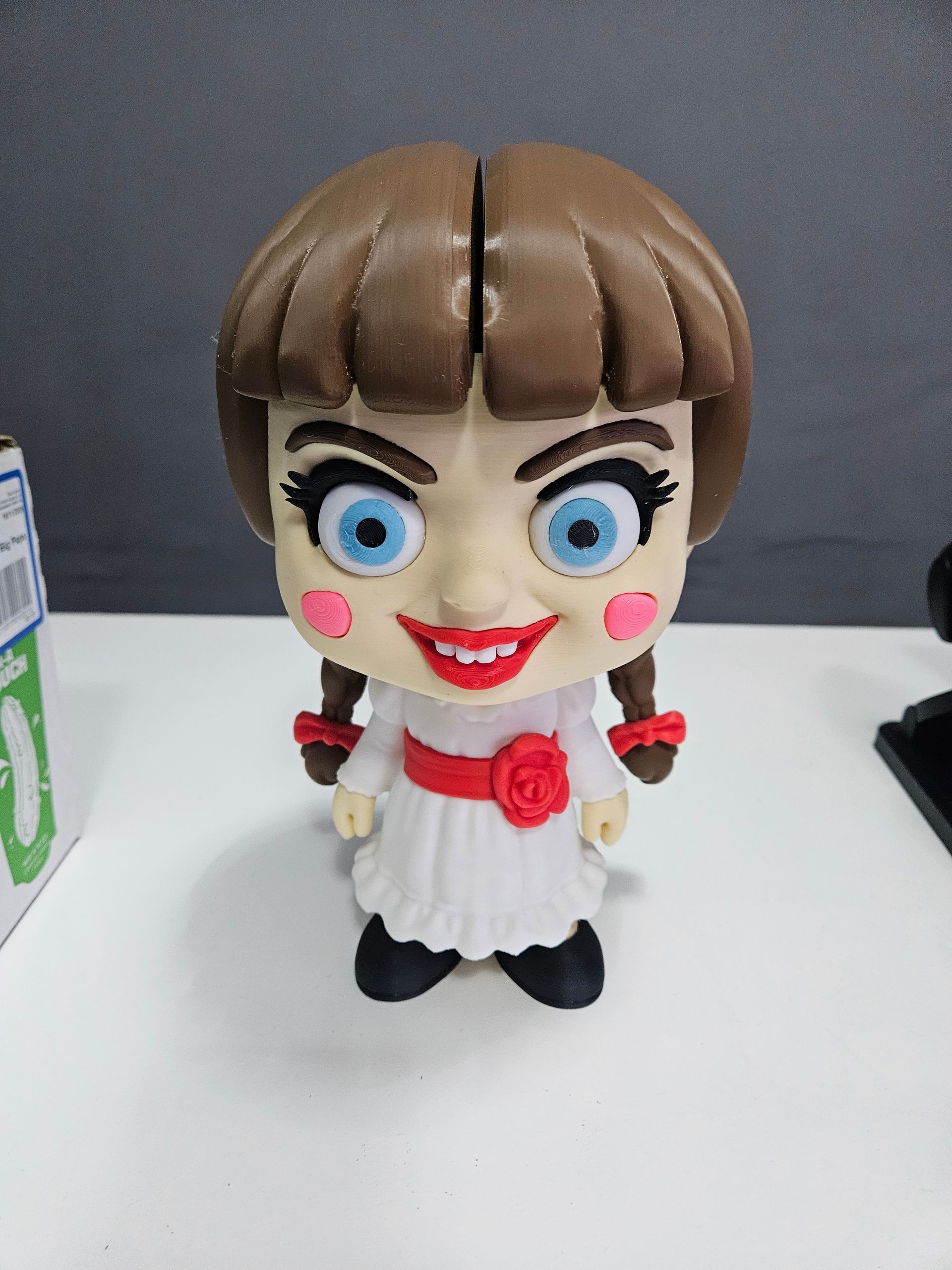 Small doll with brown bob hair, big blue eyes, red cheeks, white dress, and black shoes on a table.