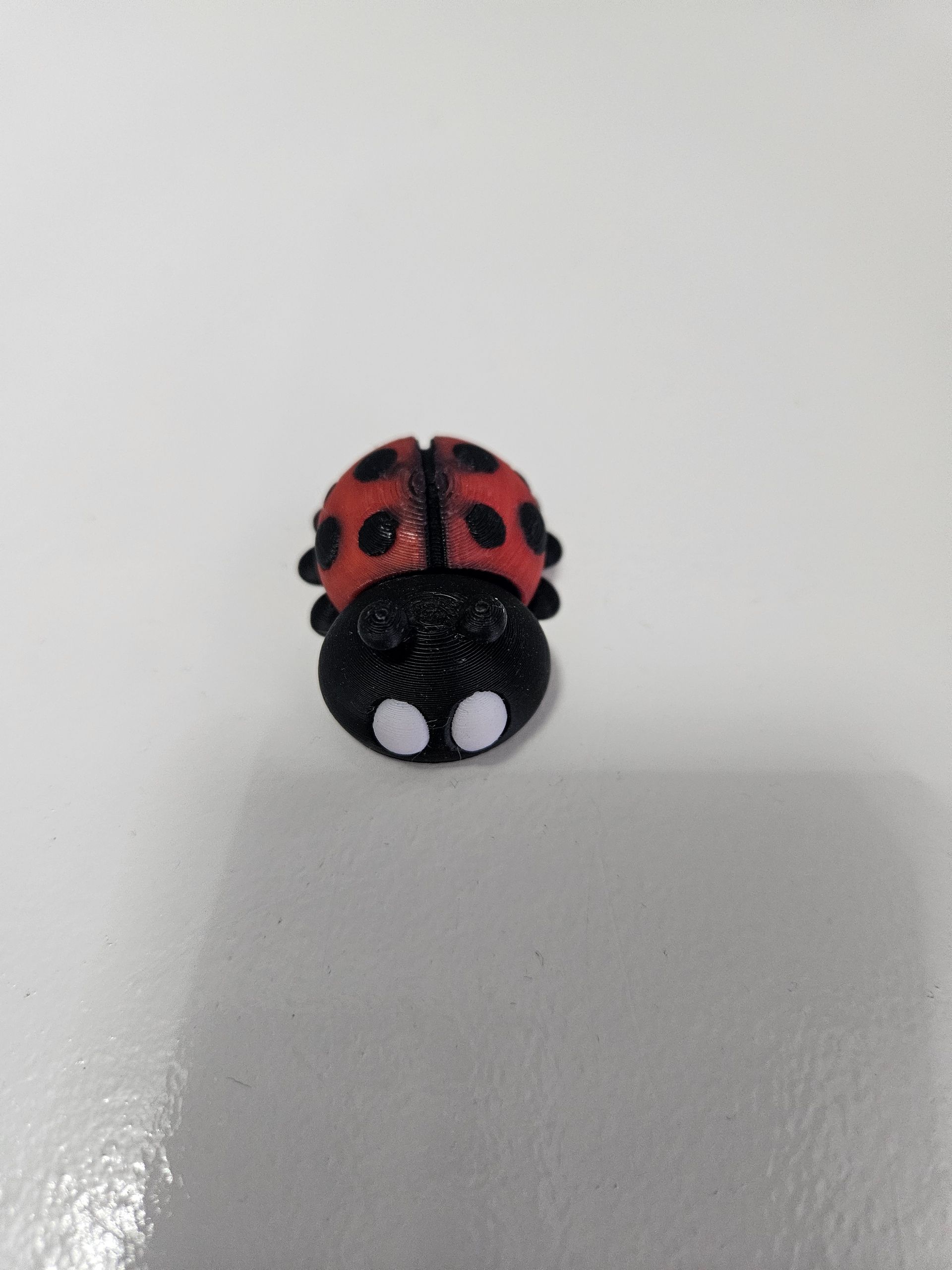 A small, beaded ladybug on a white surface; red shell with black spots and a black body with white eyes.