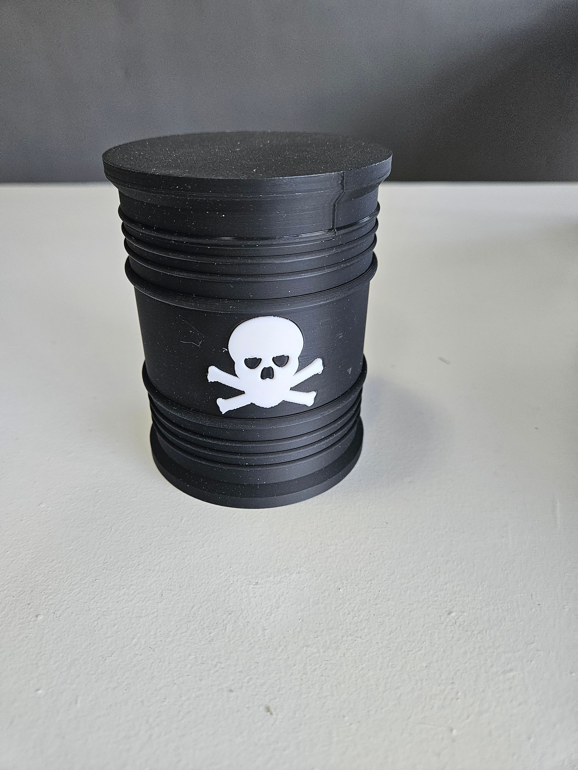 Black 3D-printed container with a skull and crossbones symbol; rests on a gray surface.