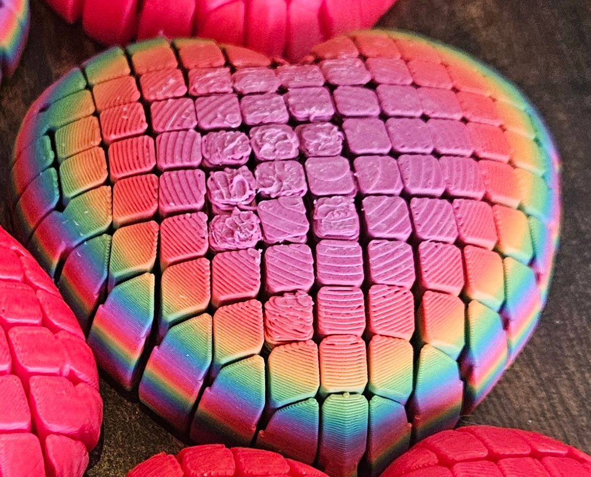 Rainbow-colored, segmented heart shape, with a grid pattern, on a dark surface, surrounded by similar objects.