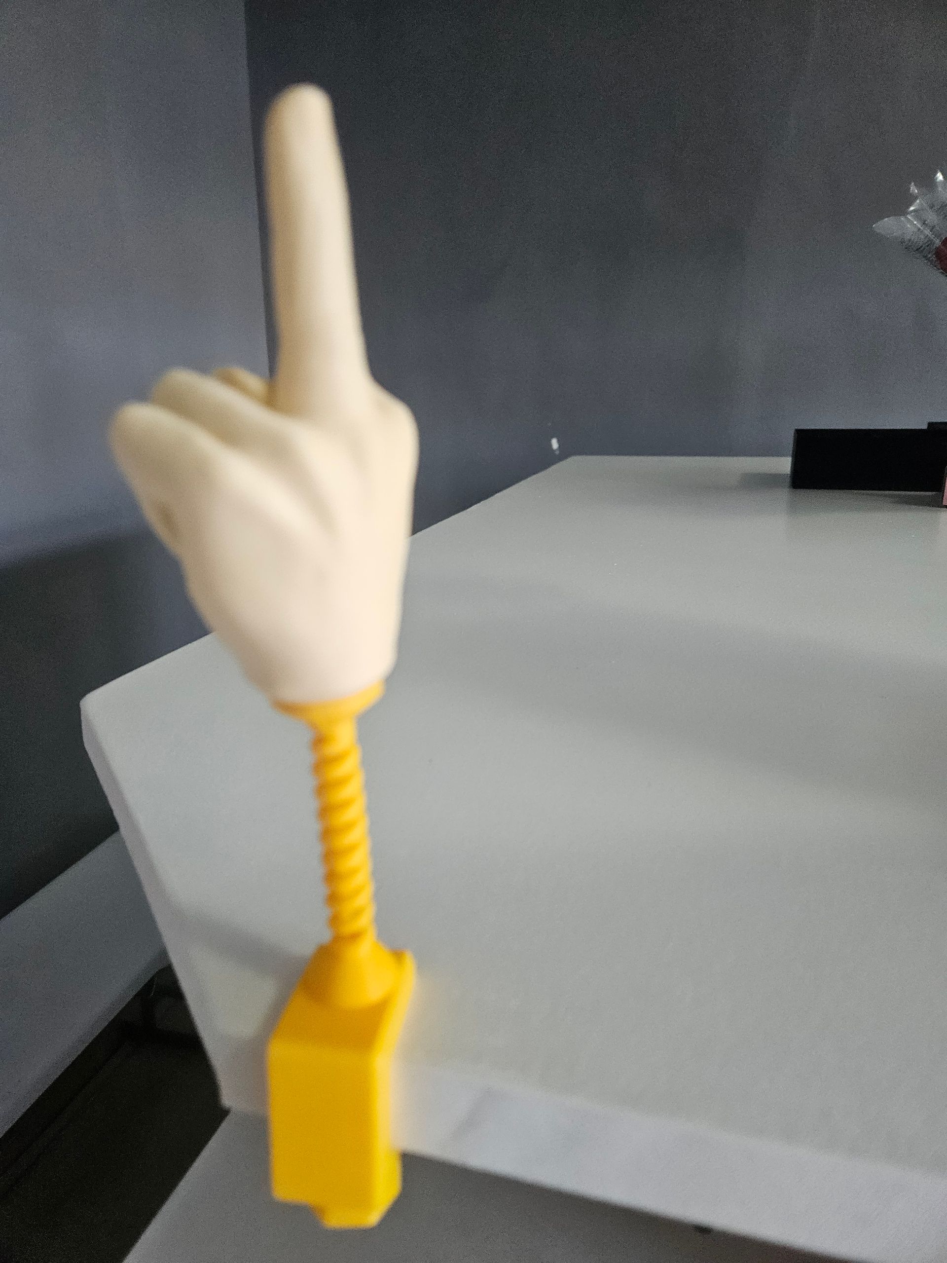 A yellow hand-shaped desk toy with its index finger pointing up. It's mounted on a yellow spring attached to a desk.