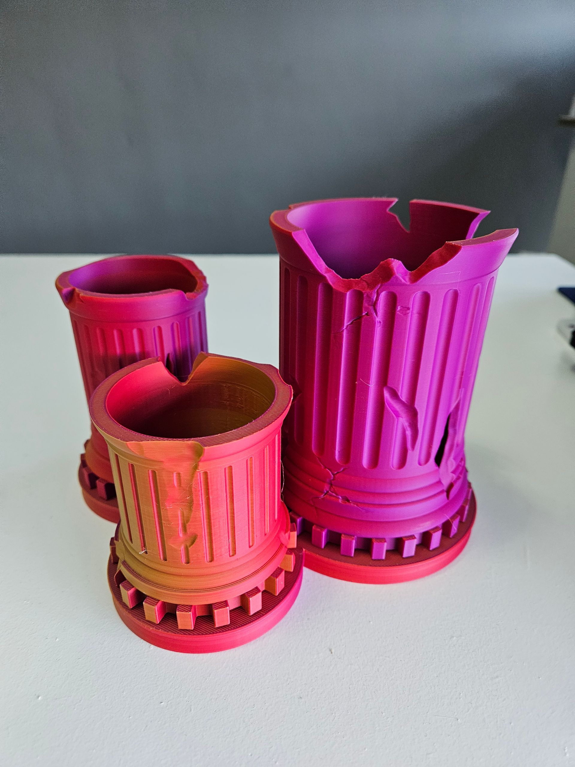Three colorful, 3D-printed, pillar-shaped containers with damaged edges. They are shaded red, orange, and purple.
