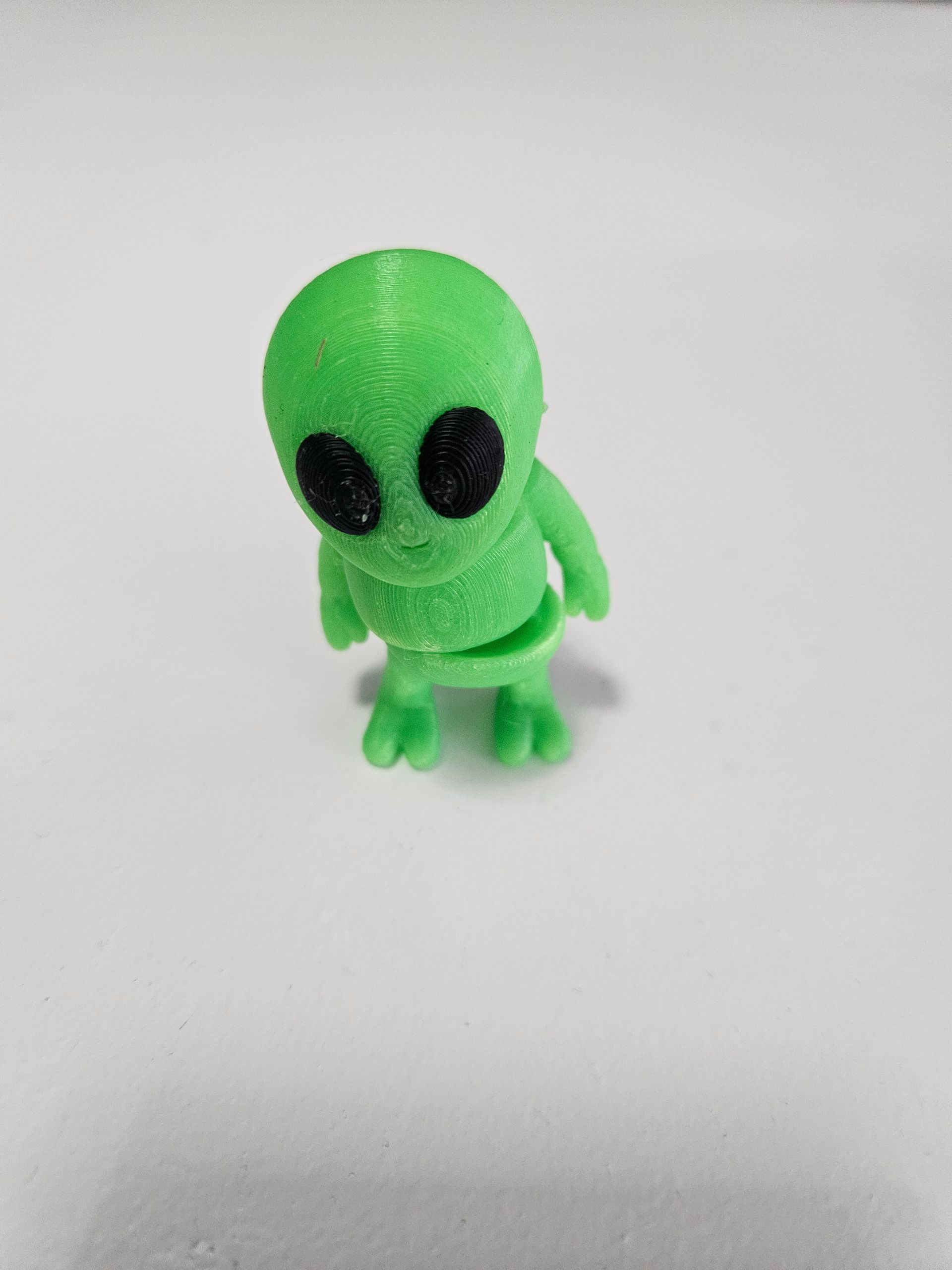 Green, 3D-printed alien figurine with large black eyes, standing on a white surface.