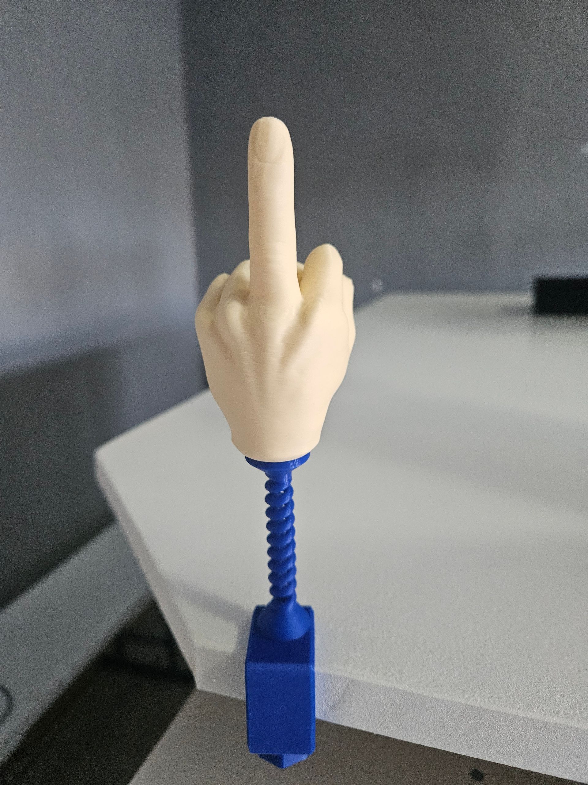 3D-printed hand giving the middle finger, cream-colored hand attached to a blue base and spring, set on a white surface.