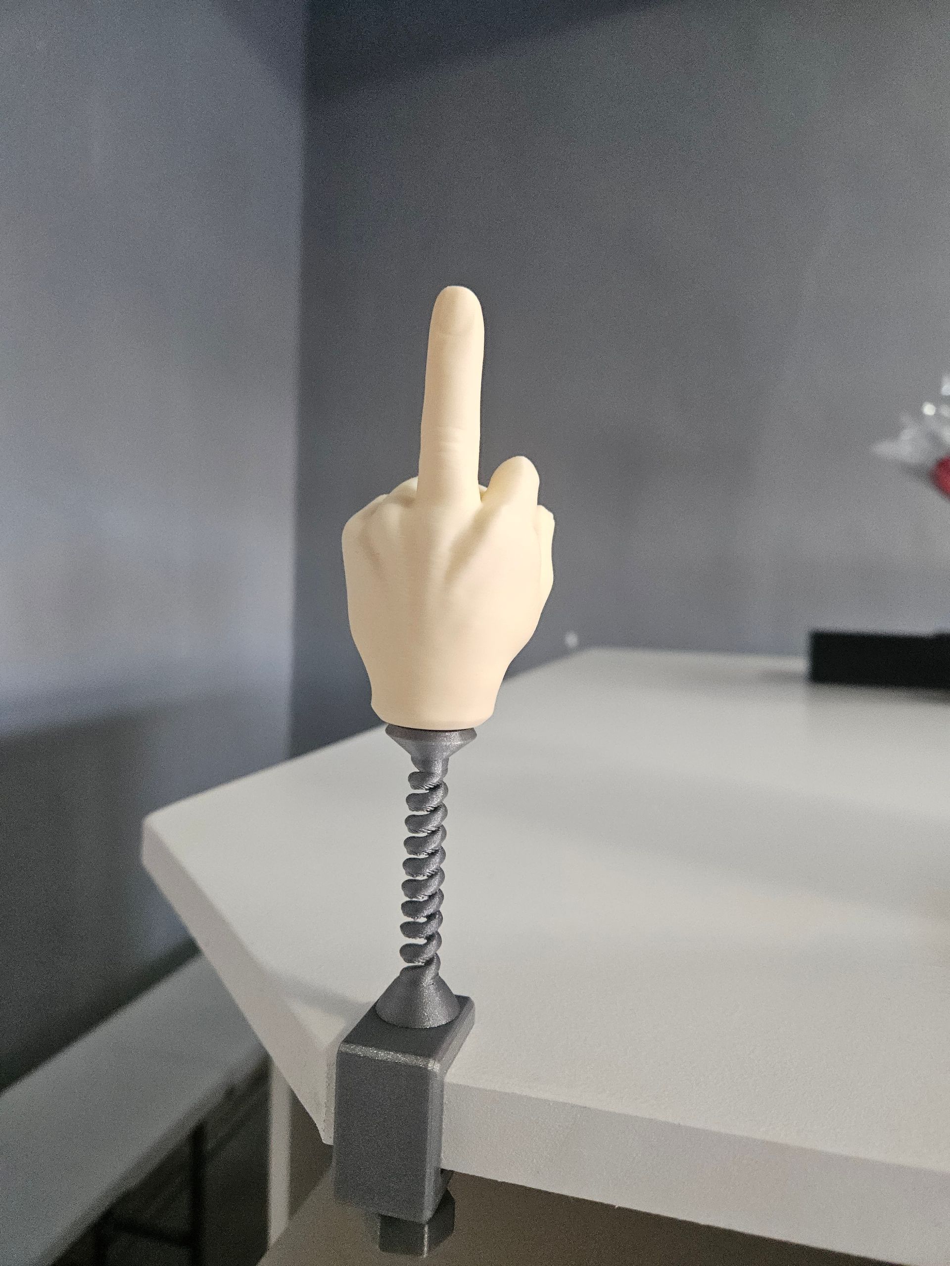 Hand figurine giving the middle finger, attached to a clamp, sitting on a white table.