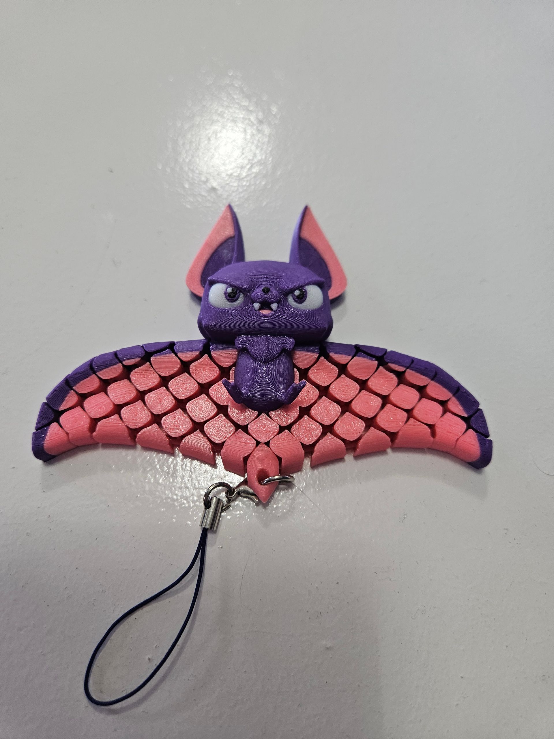 Purple and pink bat charm with an angry expression, outstretched wings, and a black cord.