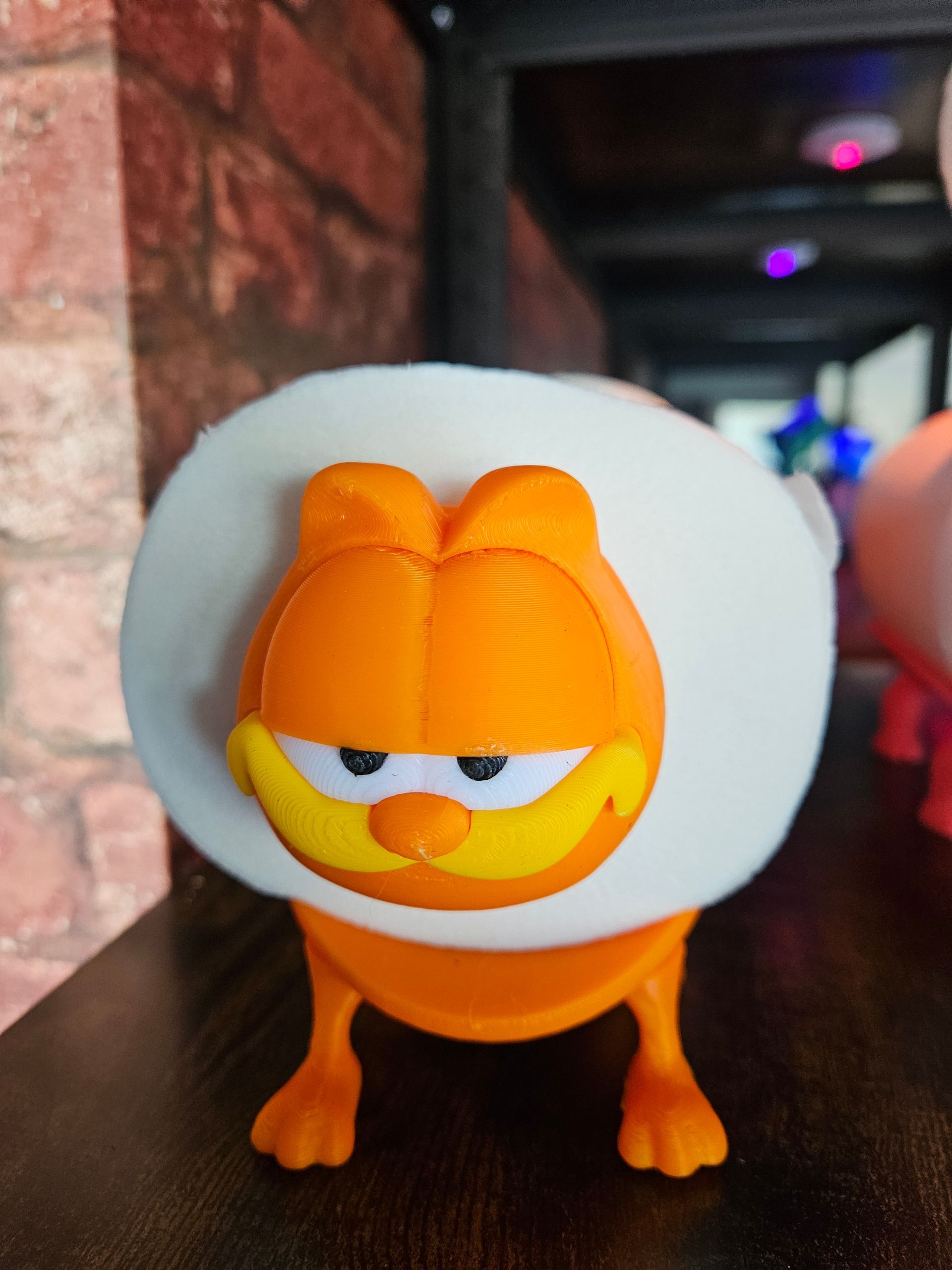 Garfield figurine with a white fluffy collar, standing indoors against a brick wall and blurred lights.