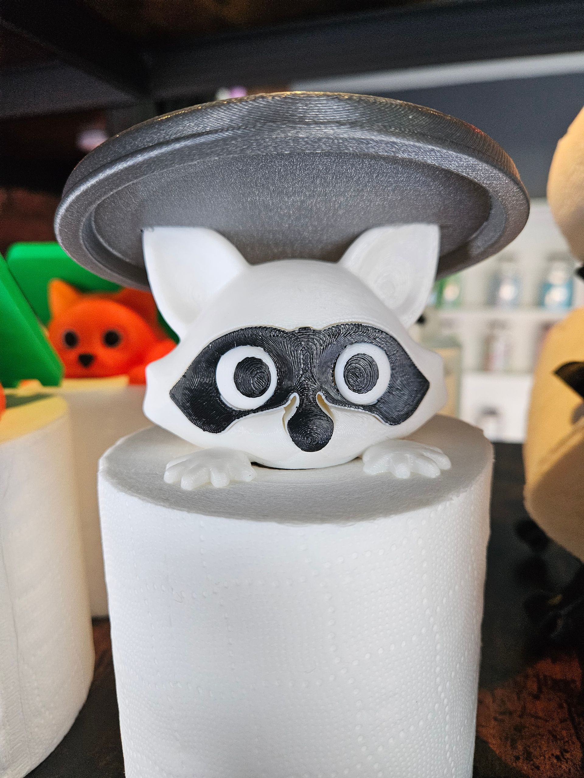 Small raccoon figurine peeking from a white cylinder under a gray felt hat, with a green and orange toy behind it.
