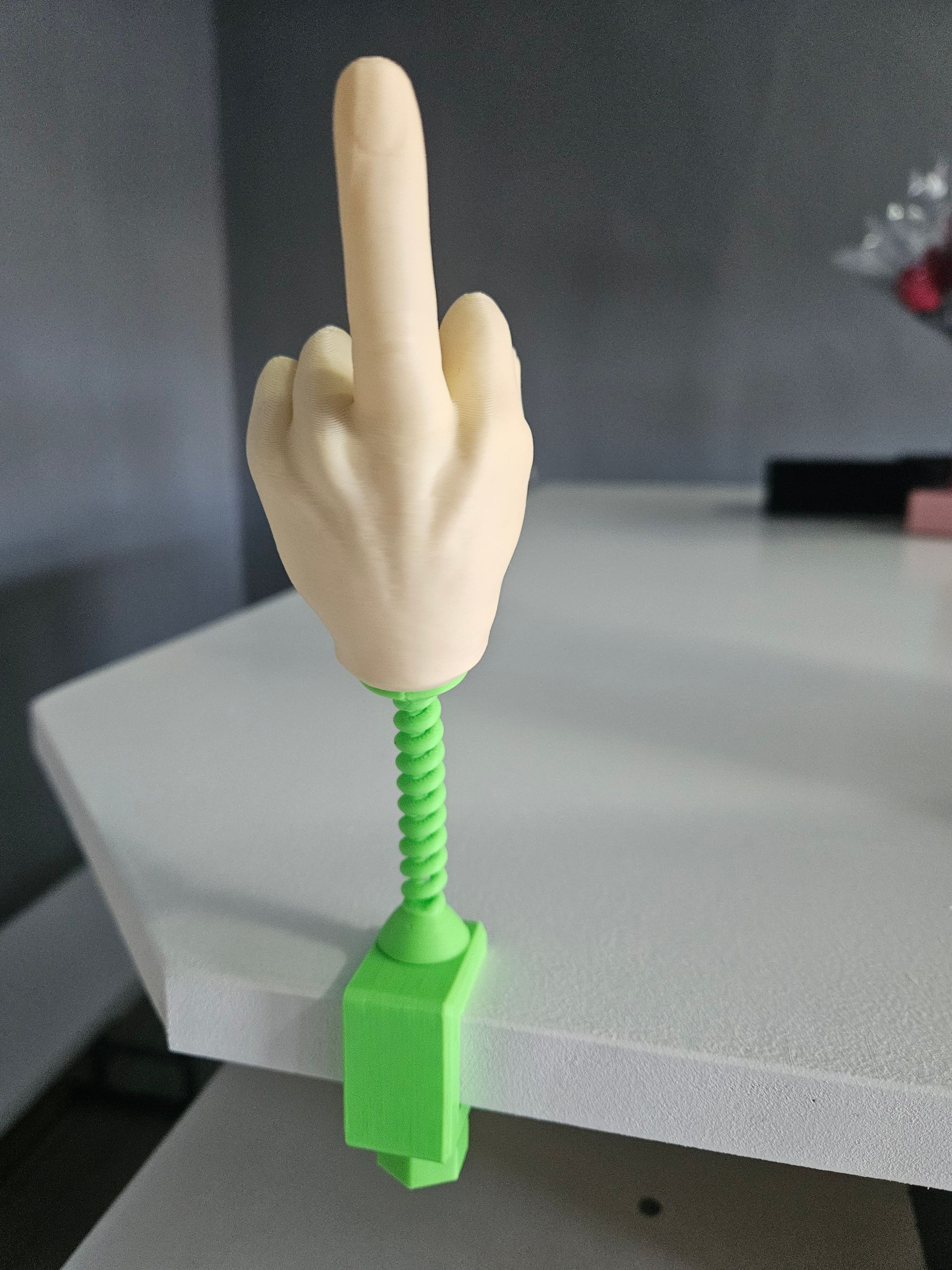 A hand giving the middle finger, mounted on a green spring and clamp, sitting on a white surface.