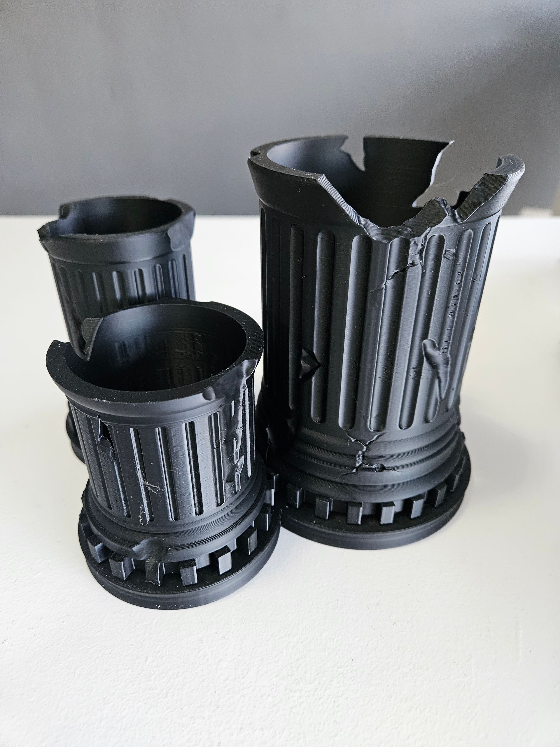 Three black, 3D-printed, pillar-shaped objects with a distressed or broken appearance, set on a white surface.
