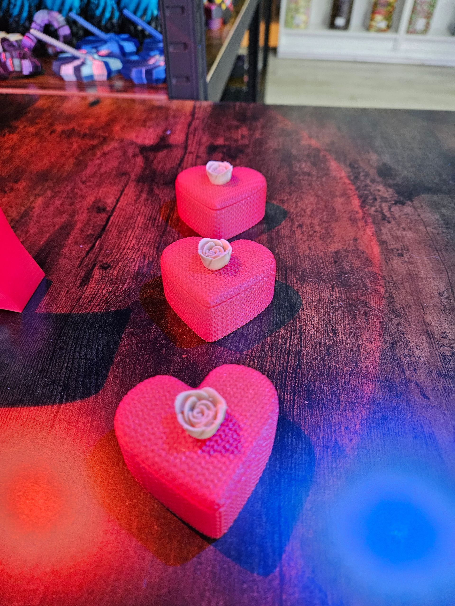 Three pink heart-shaped boxes with white rose accents on a wooden surface with red and blue lighting.