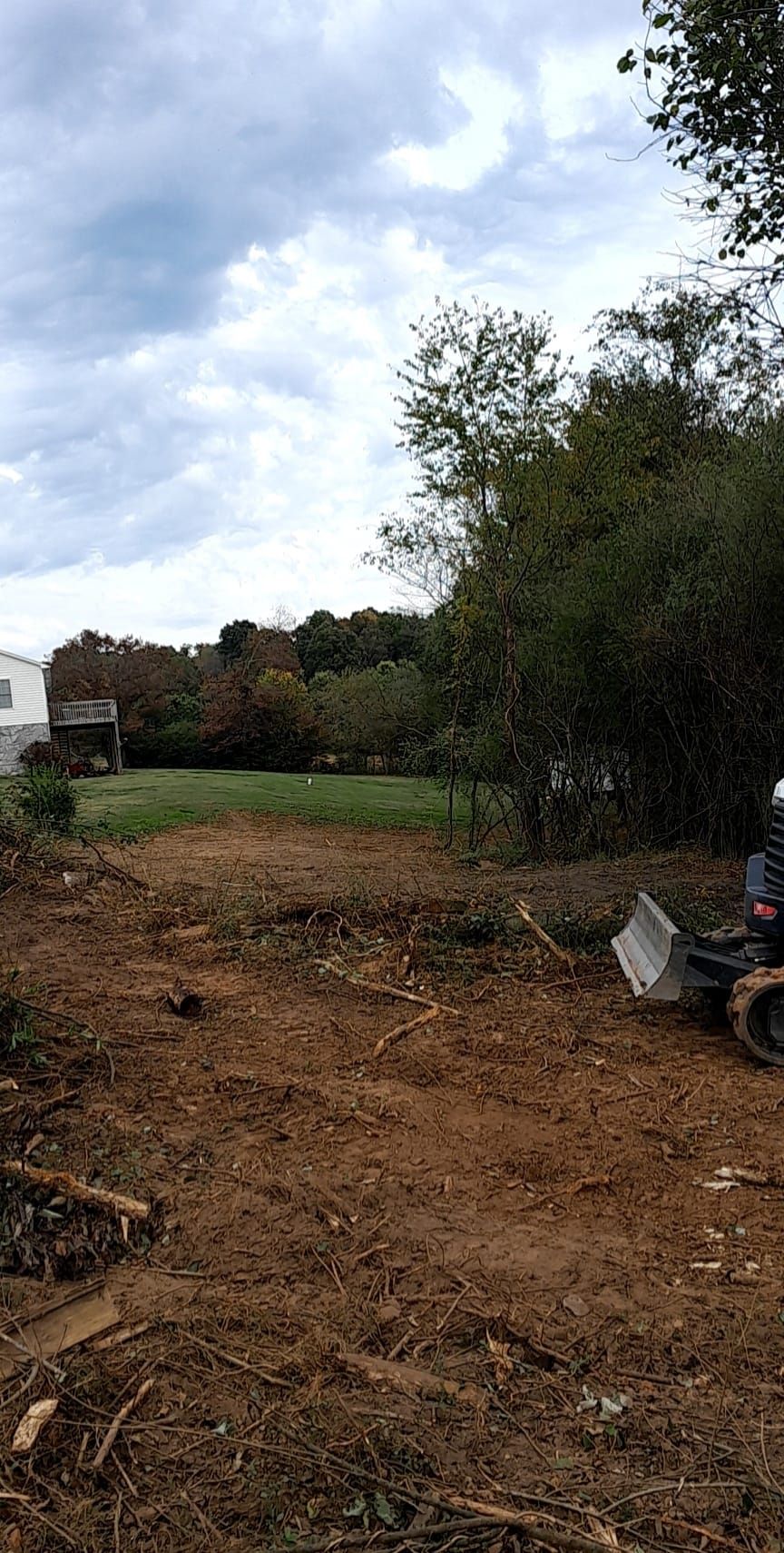 An excavator clearing a muddy lot with trees and a house in the background. Overcast sky.