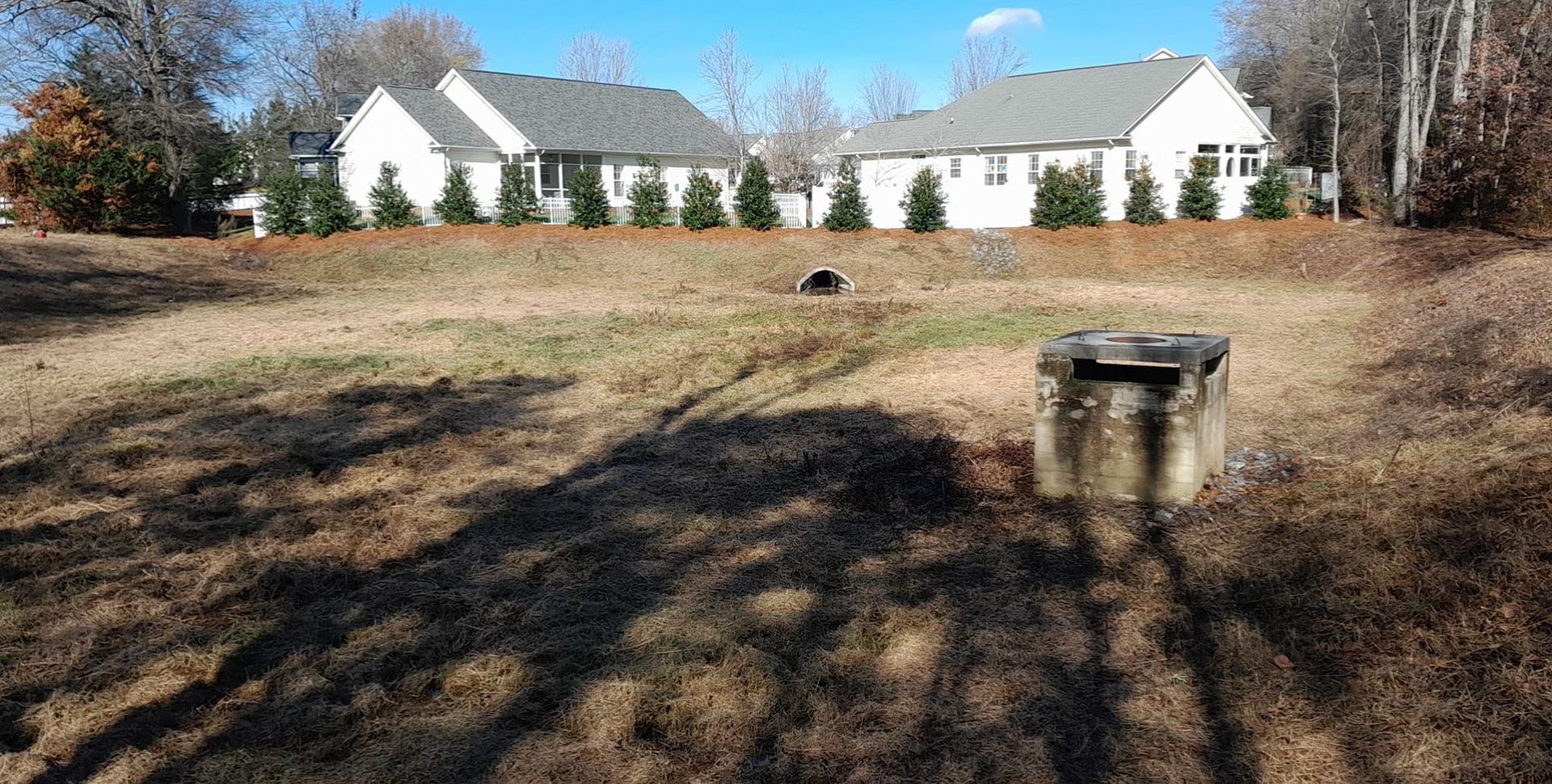 Grassy field with a concrete structure and two houses in the background on a sunny day.