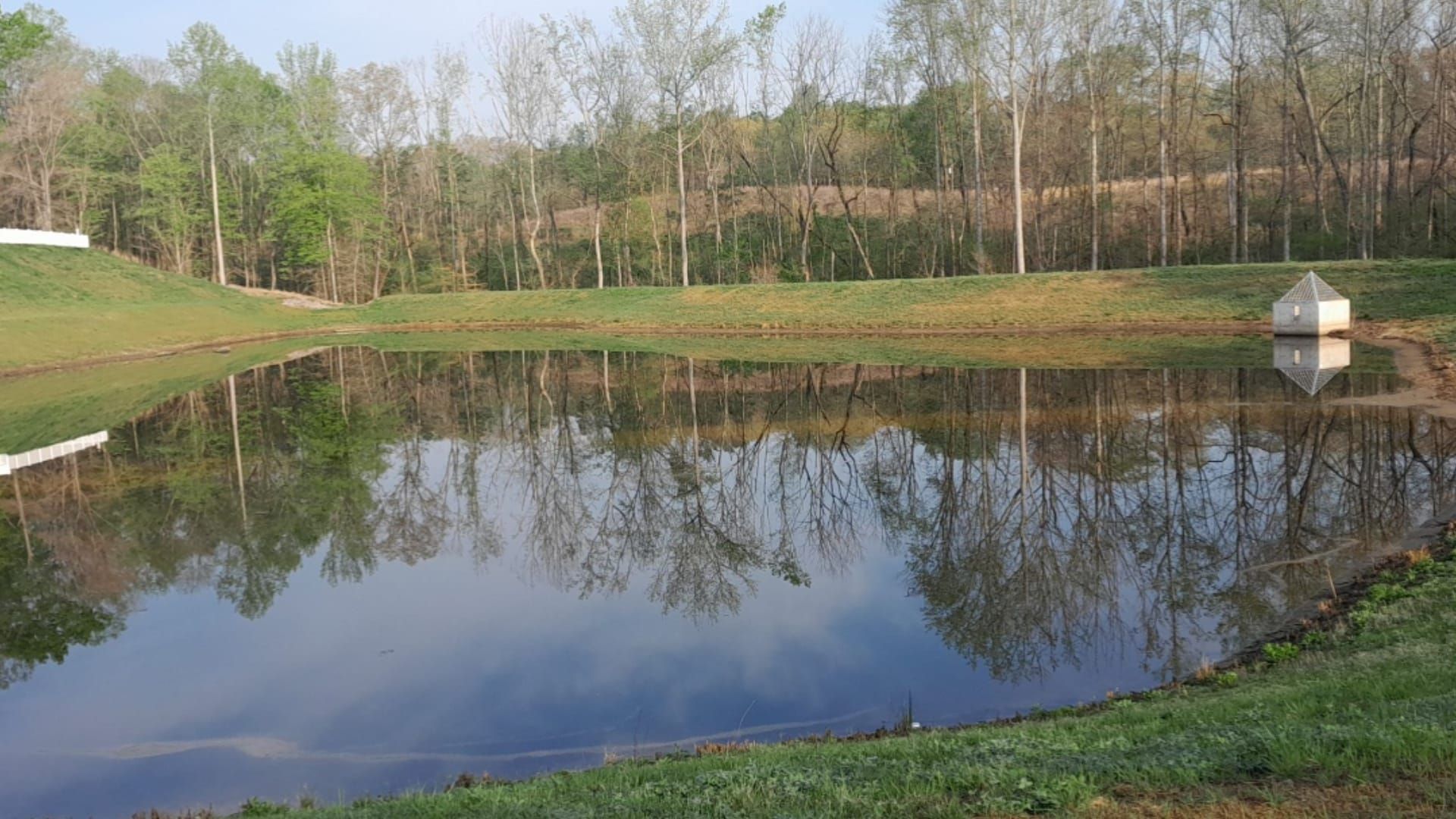 Calm pond reflects trees and sky. Small building on the right bank, grassy foreground.