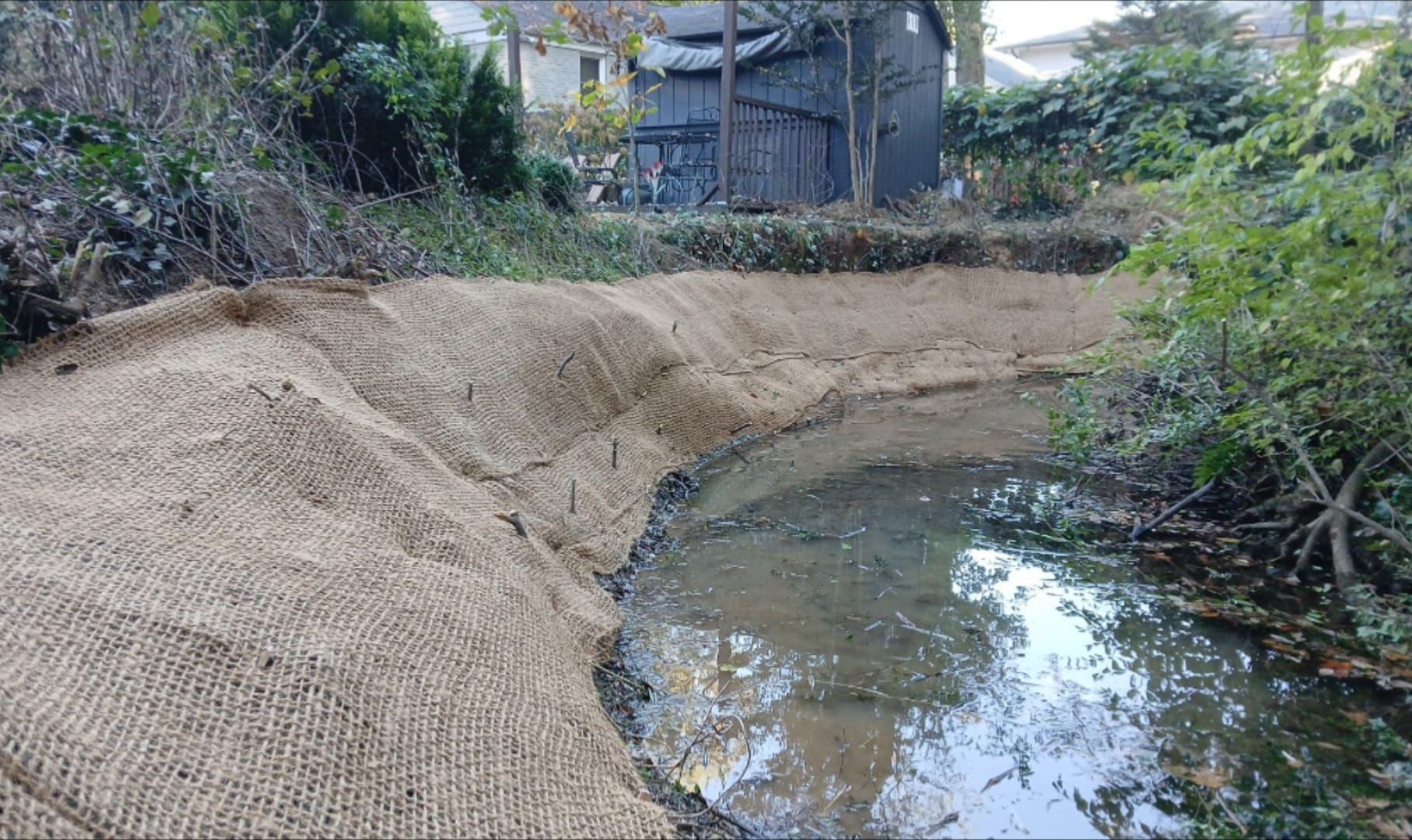 Erosion control matting lines the bank of a creek. The tan matting covers the sloped bank near the water's edge.