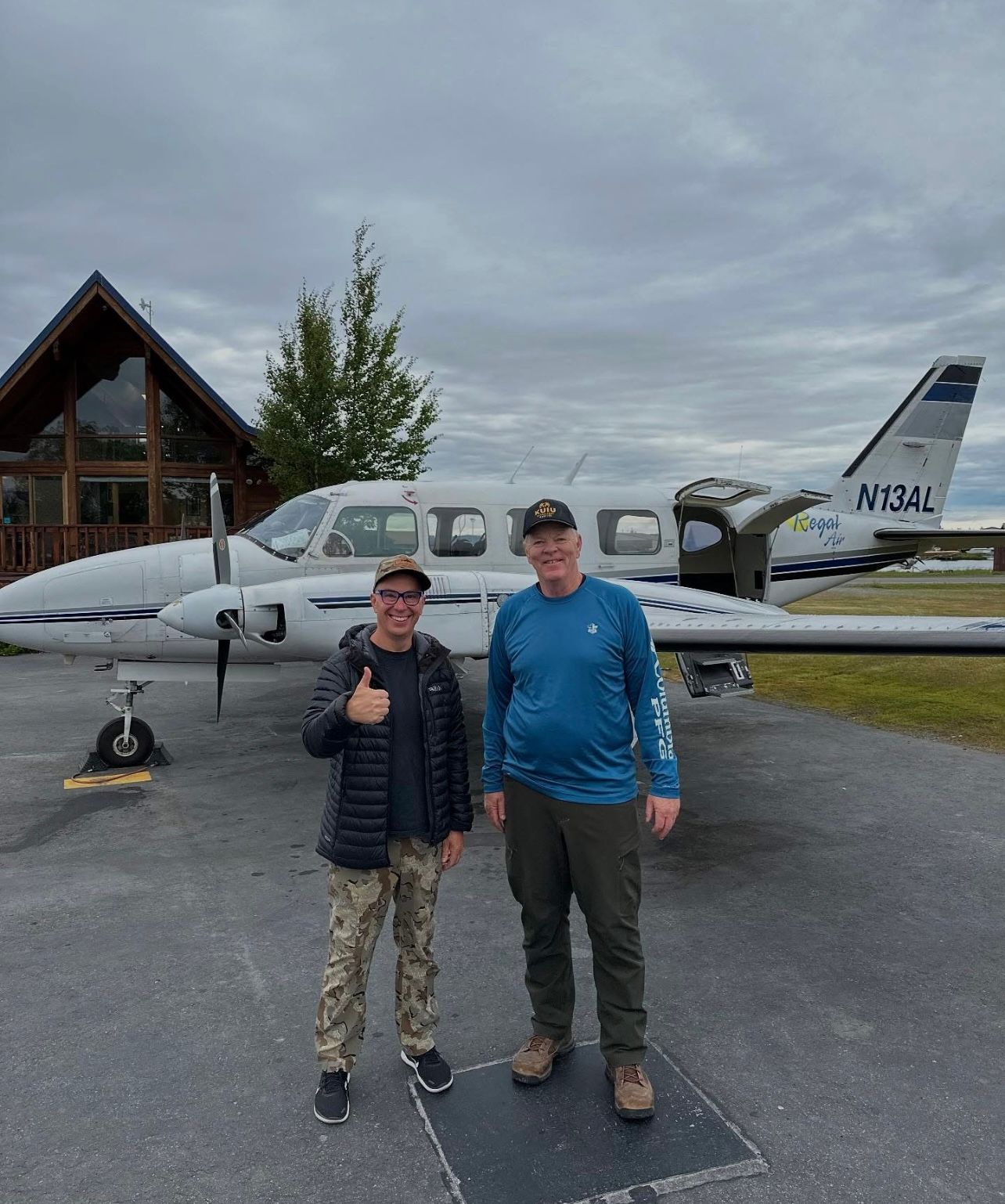 Two men stand in front of a small airplane. One gives a thumbs up. A wood-paneled building is in the background.
