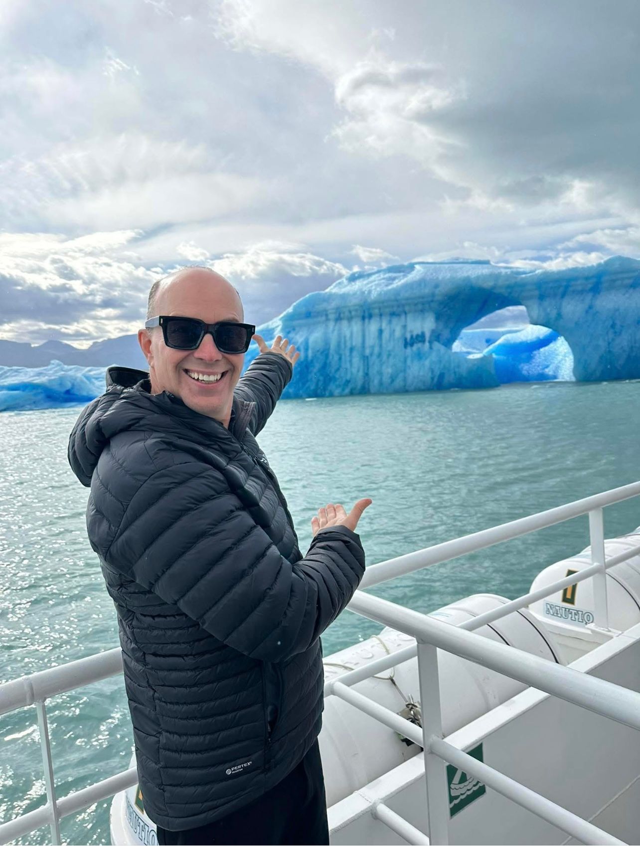 Man points toward a blue iceberg with a hole in the water from a boat, cloudy sky.