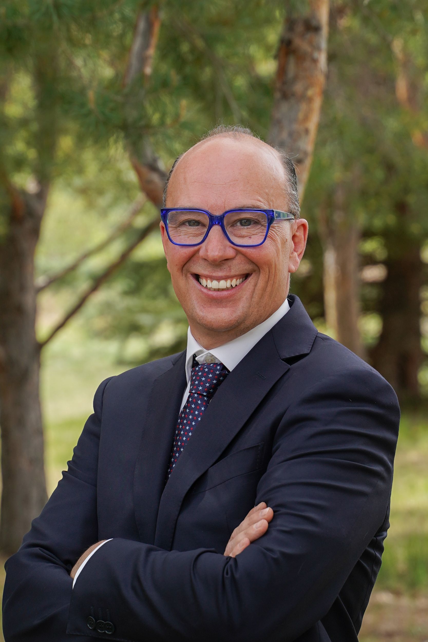Man in suit with blue glasses smiles outside with arms crossed.