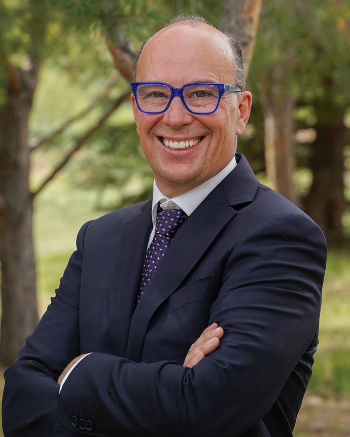 Man in suit with blue glasses smiles outside with arms crossed.