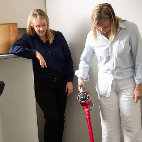 A woman is cleaning the floor with a vacuum cleaner while an occupational therapist asses her ability.