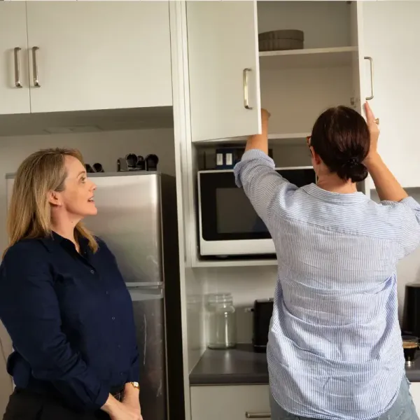 Two women are standing in a kitchen looking into a cabinet.