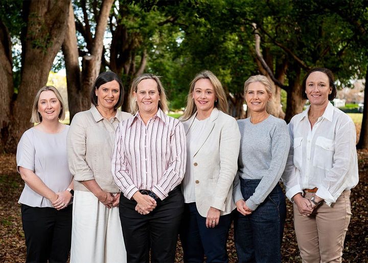 Six people standing outdoors in front of trees, smiling at the camera.