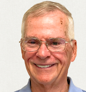 A smiling person with gray hair and glasses wearing a light blue collared shirt, against a plain white background.