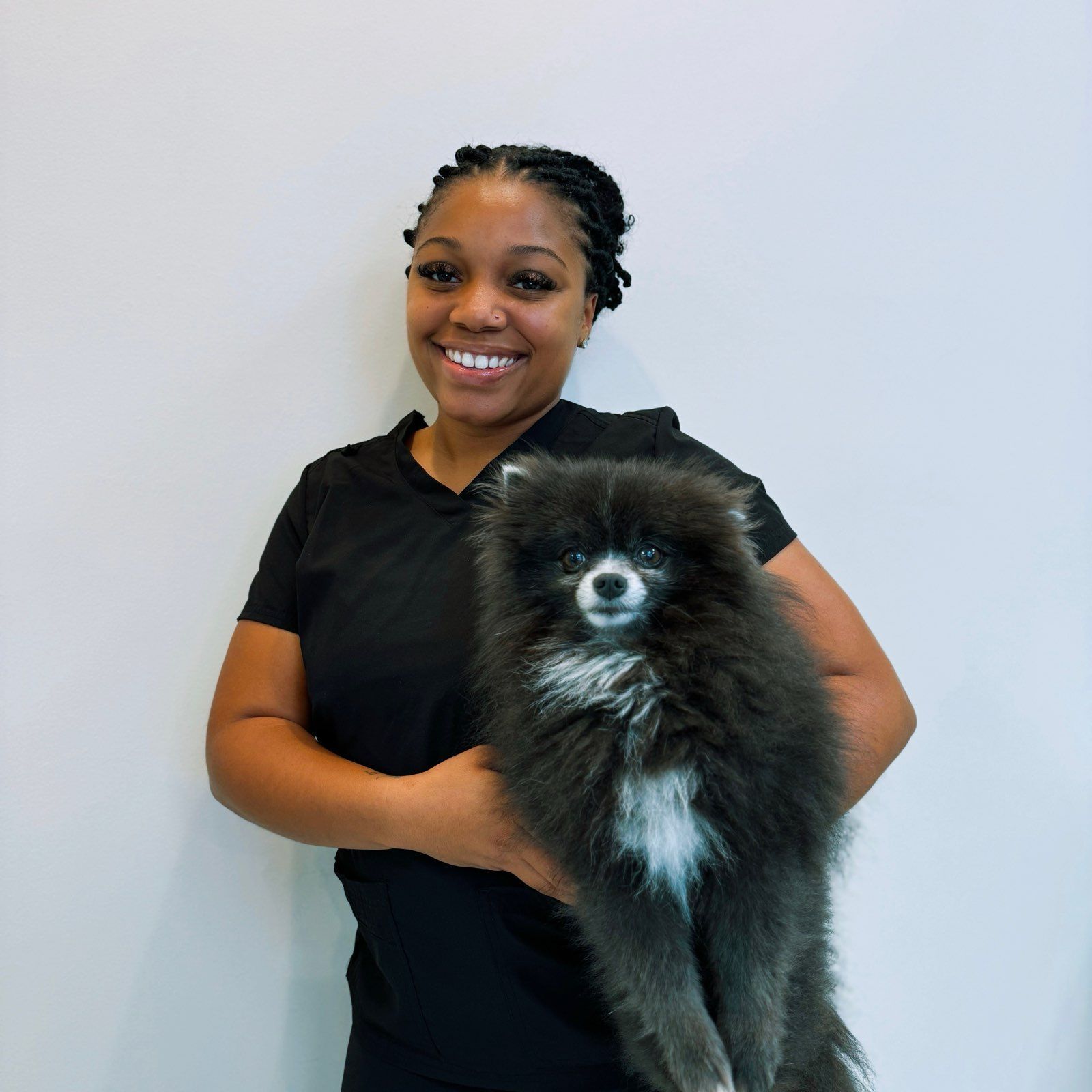A smiling person stands against a plain white background, holding a fluffy black Pomeranian dog with a white chest patch.